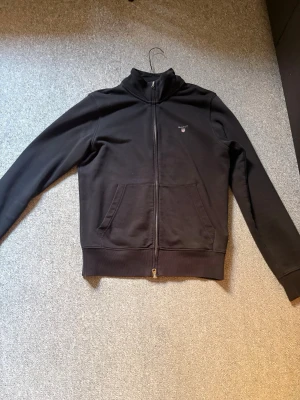 Svart zip-hoodie från GANT - Svart hoodie från GANT med dragkedja hela vägen, ribbade muddar och två stora fickor framtill. Klassisk design med liten GANT-logga på bröstet. Skön och stilren modell i mjukt material, perfekt till jeans eller joggers.