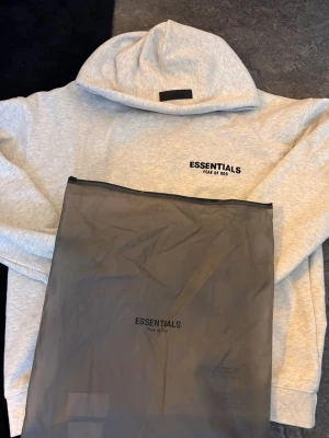 Grå hoodie Essentials Fear of God - Snygg grå hoodie från Essentials Fear of God med svart logga på bröstet och stor text på ryggen. Klassisk passform med huva och ribbade muddar. Perfekt för dig som gillar streetwear och vill ha en stilren look.