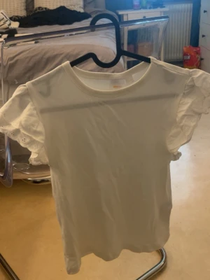 Vit topp med volangärmar från Gina Tricot - Söt vit topp från Gina Tricot med volangärmar i broderad spets. Toppen har rund halsringning och är tillverkad i mjuk bomull. Perfekt för dig som vill ha en enkel men ändå detaljerad look.
