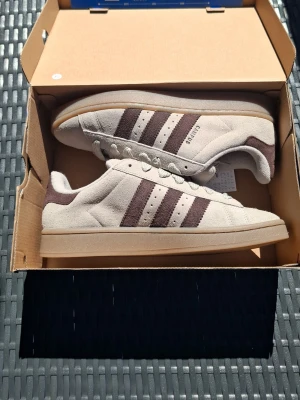 Beige Adidas sneakers med bruna ränder - Snygga beige sneakers från Adidas med klassiska tre bruna ränder på sidan och Adidas-logga på plösen. Skorna har en låg profil och är gjorda i mocka med gummisula. Perfekta för dig som gillar stilrena och tidlösa sneakers. SKORNA KOMMER MED BOX OCH ETT EXTRA PAR BRUNA SNÖREN