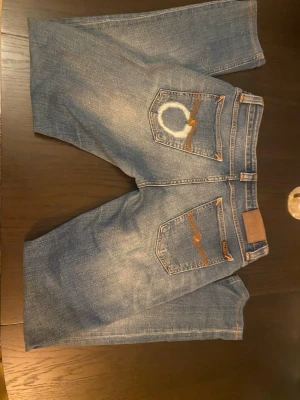 Blå raka jeans med kontrastsömmar - Säljer ett par blå jeans med raka ben och tydliga kontrastsömmar. Jeansen har slitningar och en unik vit cirkeldetalj på ena bakfickan. Klassisk femficksmodell och knappgylf. Materialet är denim i bomull.