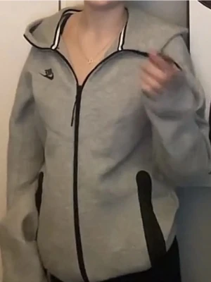 Grå zip hoodie från Nike - Säljer en grå hoodie från Nike med dragkedja framtill och svart logga på bröstet. Hoodien har svarta detaljer vid fickorna och en stor huva. Perfekt för en avslappnad och sportig stil.