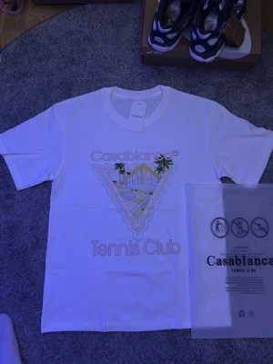 Vit Casablanca Tennis Club t-shirt - Snygg vit t-shirt från Casablanca med färgglatt tryck framtill som visar palmer och tennisbana samt texten 'Casablanca Tennis Club'. Sköna färger och allmänt clean tröja, fraktar samma dag!
