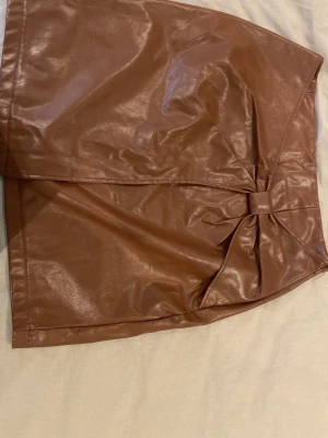 Brun kortkjol i skinnimitation med rosett - Snygg brun kortkjol i skinnimitation med en dekorativ rosettdetalj framtill. Kjolen har en slät yta och enkel design som ger en trendig look. Skriv till mig ifall du undrar mått🥰🤎