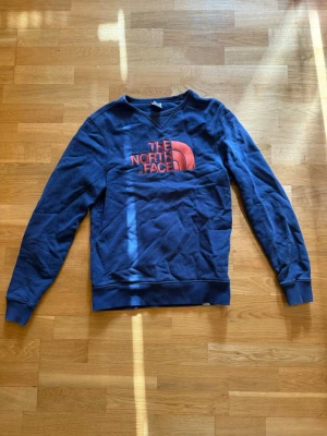Blå sweatshirt från The North Face - Mörkblå sweatshirt från The North Face med orange broderad logga på bröstet. Tröjan har rund halsringning, ribbade muddar och är tillverkad i mjuk bomullsblandning. Perfekt för en avslappnad och sportig stil.
