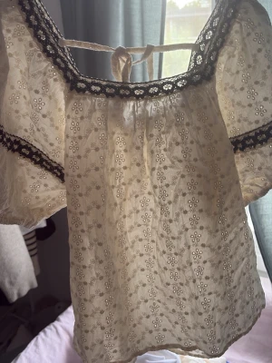 Vit blus med spets och broderi - Romantisk vit blus med broderade blommor och svarta spetsdetaljer vid halsringning och ärmar. Puffiga korta ärmar och knytband i nacken ger en söt vintagekänsla. Perfekt för dig som gillar boho-stil.