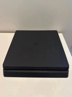 PlayStation 4 Slim svart spelkonsol - Säljer en svart PlayStation 4 Slim spelkonsol från Sony. Konsolen har en stilren, tunn och fyrkantig design med mattsvart finish och PlayStation-loggan på ovansidan. Perfekt för gaming och streaming av filmer och serier.