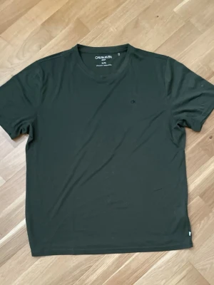 Mörkgrön t-shirt från Calvin Klein - En stilren mörkgrön t-shirt från Calvin Klein med diskret 'ck'-logga på bröstet. Klassisk rund halsringning och korta ärmar. Tillverkad i mjuk bomull som känns skön mot huden. Perfekt basplagg för en clean och enkel look.
