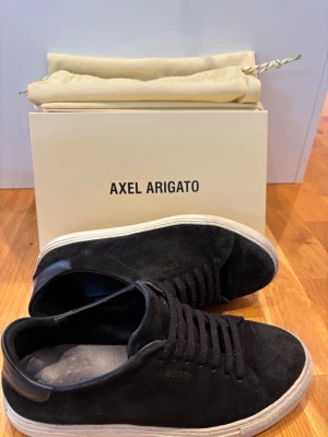 Svarta sneakers från Axel Arigato - Stilrena svarta sneakers i mocka från Axel Arigato med vit sula och svarta skosnören. Låg modell med diskret logga på sidan och detaljer i läder vid hälen. Perfekta för en clean och modern look. Använts i en säsong, skolåda och skopåse på köpet