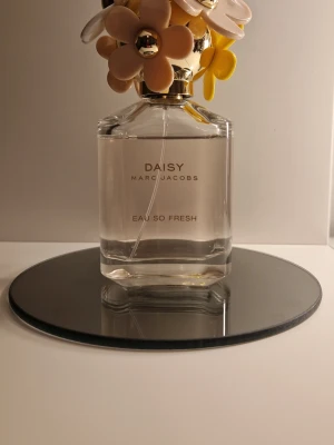 Daisy Eau So Fresh parfym - Daisy Eau So Fresh från Marc Jacobs är en parfym i en genomskinlig glasflaska med guldtext. Locket är dekorerat med stora blommor i pastellfärger som gult, beige och vitt. Flaskan har en elegant och lekfull design som sticker ut på hyllan.