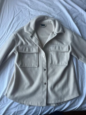 Beige overshirt med stora fickor - Overshirt i beige med två stora bröstfickor och tryckknappar framtill. 