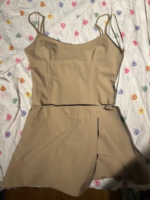Beige jumpsuit/klänning med tunna axelband - Trendig beige jumpsuit med tunna axelband och subtila vertikala ränder. Modellen har en omlottkjol över shortsdelen och markerad midja för en snygg siluett. Tillverkad i ett mjukt, vävt tyg som ger en clean och modern look. Perfekt för dig som gillar stilrena plagg med twist.