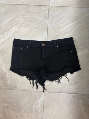 Svarta jeansshorts med fransig kant - Snygga svarta jeansshorts med rå, fransig kant och klassisk femficksdesign. Shortsen har låg midja och knappgylf. Perfekta för varma dagar och ger en cool, avslappnad vibe.