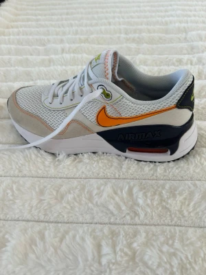 Nike Air Max vita sneakers - Nike Air Max sneakers med vit mesh och beige mockadetaljer. Orange Swoosh på sidan, svarta och vita detaljer runt sulan samt Air Max-logo. Skorna har vita snören och gröna accenter på plösen. Perfekt för dig som gillar sportig och trendig stil. Skon är 24 centimeter. Skorna är nya.