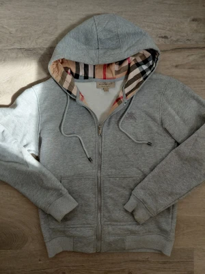 Grå zip hoodie från Burberry - Snygg grå zip hoodie från Burberry med klassiskt rutigt foder i huvan och broderad logga på bröstet. Mjuk bomullsblandning, dragsko i huvan och två fickor framtill. Perfekt för en avslappnad och stilren look.