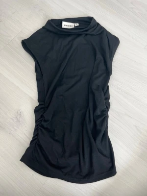 Svart ärmlös topp från Kappahl - Svart ärmlös topp från Kappahl med hög krage och snygg rynkning på sidan. Tillverkad i mjukt material som känns skönt mot huden. Perfekt att styla med jeans eller kjol för en clean look.