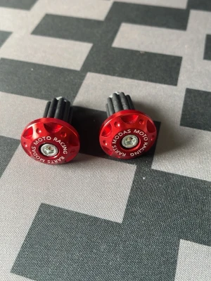 Röda styrändar till moped - Säljer ett par röda styrändar i metall med texten 'MOTO RACING PARTS MODAS' runt kanten. De har en rund form och svart plastdetalj som fästs i styret. Perfekt för dig som vill ge hojen en sportig look. Aldrig använt!!!!