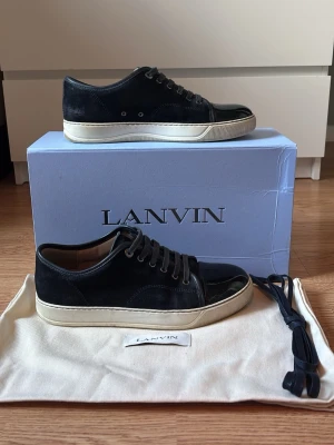  Lanvin sneakers i mocka och lack - Snygga svarta sneakers från Lanvin med ovandel i mocka och lackad tå. Skorna har vita sulor, svarta skosnören och diskreta metalldetaljer vid snörningen. Klassisk låg modell som passar till det mesta. Extra skosnören och dustbag medföljer.