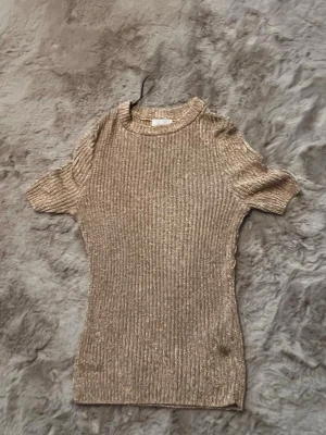 Ribbad beige topp med glitter - Säljer en ribbad topp i beige med glittriga inslag. Toppen har korta ärmar och rund halsringning. Materialet känns mjukt och stretchigt, perfekt för dig som vill ha något enkelt men ändå lite extra.