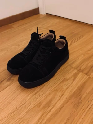 Svarta loubs i sammet - Snygga nya svarta sneakers som använts några gånger. De är i sammet med röd sula som går att tvätta enkelt och svarta skosnören. På hälen finns en vit broderad text som ger en unik touch. Insidan är beige och skon har en klassisk låg profil som passar till många stilar.