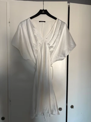 Vit omlottklänning i satin - Säljer en elegant vit kortklänning i satin med omlottdesign och v-ringning. Klänningen har vida, korta ärmar och ett mjukt, glansigt material som ger ett lyxigt intryck. Perfekt för dig som vill ha en stilren och trendig look.