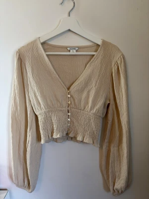 Beige blus med knappar från Monkl - Säljer en supersöt beige blus från Monkl med långärmad design, v-ringning och dekorativa pärlknappar framtill. Blusen har smockad midja och volangkant nedtill, tillverkad i ett lätt, krinklat tyg som ger en avslappnad men trendig look.
