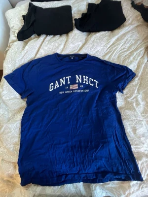 Blå GANT t-shirt med tryck - Snygg blå t-shirt från GANT med vit text och tryck på bröstet där det står 'GANT NHCT New Haven Connecticut'. Klassisk passform och rund halsringning. Perfekt till jeans eller shorts för en avslappnad look.
