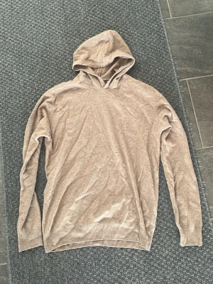 Beige hoodie i mjukt material från Oscar Jacobsson  - Säljer en stilren beige hoodie med huva och långa ärmar. Hoodien är tillverkad i ett mjukt och tunt material, perfekt för en avslappnad look. Ribbstickade muddar vid ärmslut och nederkant ger en snygg passform.