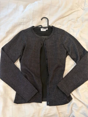 Glittrig svart kofta från Saint - Snygg svart kofta från Saint med diskret glittrigt material. Rund halsringning, långa ärmar och knäppning framtill. Perfekt för dig som vill addera lite extra till din outfit. Enkel och stilren design som passar till mycket.