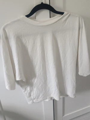 Vit trekvartsärm t-shirt i bomull - En basic vit t-shirt med trekvartsärm och rund hals. Tillverkad i mjuk bomull för en avslappnad och enkel look. Perfekt att matcha med jeans eller kjol för en clean stil.
