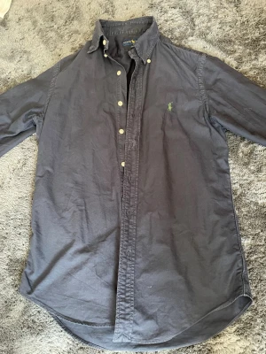 Mörkblå skjorta från Ralph Lauren - Klassisk mörkblå skjorta från Ralph Lauren med button-down krage och långa ärmar. Skjortan har vita knappar och den ikoniska gröna logotypen broderad på bröstet. Tillverkad i mjukt bomullstyg och har rundad nederkant.