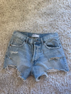 Ljusa denimshorts med slitningar från Zara - Säljer ett par ljusblå denimshorts från Zara med hög midja och råa, fransiga kanter. Shortsen har flera slitningar framtill och klassisk femficksdesign. Perfekta för sommaren och ger en avslappnad vibe.