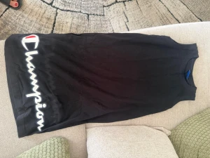 Svart ärmlös linnetopp Champion - Svart ärmlös linnetopp från Champion med stort vitt och rött logotryck längs sidan framtill. Toppen har rund halsringning och är gjord i mjuk bomull, perfekt för en sportig och avslappnad stil.