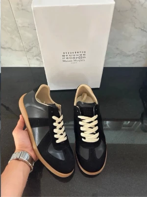 Maison Margiela svarta sneakers - Maison Margiela sneakers i svart läder och mocka med vita snören och beige gummisula. Klassisk låg modell med stilren design och diskreta detaljer. Perfekta för dig som gillar minimalistisk och trendig stil.