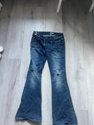 Blå bootcut jeans med slitningar - Säljer ett par blå bootcut jeans med slitningar på båda knäna och fickor fram. Jeansen har dubbla knappar i midjan och en klassisk tvättad look. Perfekta för dig som gillar en avslappnad och trendig stil.