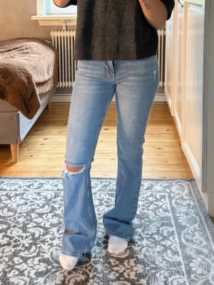 Ljusblå bootcut jeans med slitning - Säljer ett par ljusblå bootcut jeans med en snygg slitning vid knät. Jeansen är midwaist och används inte längre.