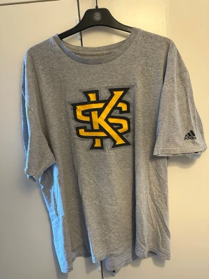 Grå Adidas t-shirt med gul logga - Säljer en grå t-shirt från Adidas med stor gul och svart logga på bröstet och Adidas-märke på ärmen. T-shirten har rund hals och är kortärmad, perfekt för en avslappnad stil. Tillverkad i mjuk bomull för skön känsla hela dagen.