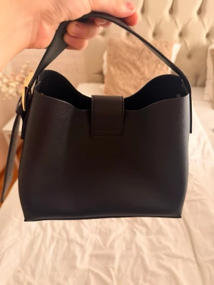 Svart bucket handväska i läderlook - Stilren svart bucket handväska i slät läderimitation med justerbart axelband och guldfärgat spänne. Väskan har en enkel men modern design med vågig kant upptill och praktiskt magnetlås. Perfekt för dig som gillar minimalistisk stil.