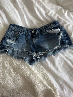 Blå slitna jeansshorts med fransar - Säljer ett par blå jeansshorts med slitna detaljer och fransig kant. Shortsen har fem fickor, bälteshällor och knappstängning. Perfekta för sommaren och ger en avslappnad vibe.