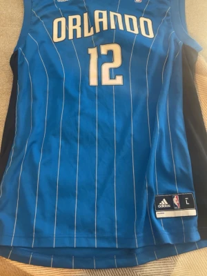 Orlando Magic basketlinne blå Adidas - Snyggt blått basketlinne från Orlando Magic med vita ränder och nummer 12, samt Howard på ryggen. Tillverkat i lätt och ventilerande material, perfekt för basket eller träning. Klassisk NBA-design med logga och Adidas-märke på nederkanten.