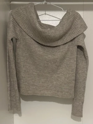 Ljusgrå ribbstickad tröja  - Säljer en ljusgrå ribbstickad tröja med lång ärm och bred, nedvikt krage som ger en snygg off-shoulder look. Tröjan är mjuk och har en rak passform, perfekt för lager på lager. Materialet ser ut att vara en ull- eller akrylblandning.