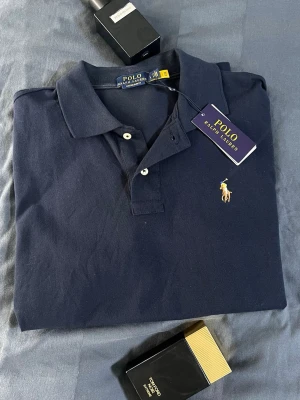 Mörkblå piké från Polo Ralph Lauren - Klassisk mörkblå pikétröja från Polo Ralph Lauren med den ikoniska broderade loggan på bröstet. Tröjan har krage, knappar framtill och är tillverkad i mjuk bomull. Perfekt för en clean och stilren look.