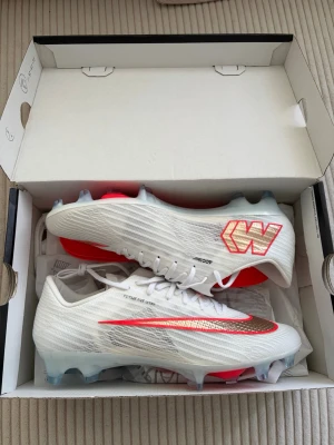 Vita Nike Mercurial fotbollsskor - Säljer ett par vita Nike Mercurial fotbollsskor med röda och guldiga detaljer. Skorna har snörning, låg profil och dobbar för spel på gräs. Tillverkade i syntetmaterial för lätt vikt och bra bollkontroll. Perfekta för dig som vill ha speed och stil på planen.