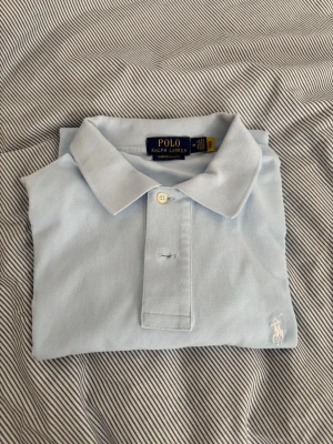 Ljusblå pikétröja från Polo Ralph Lauren - Klassisk ljusblå pikétröja från Polo Ralph Lauren med korta ärmar och vit broderad logga på bröstet. Tröjan har krage och knappar framtill, perfekt för en clean och stilren look. Storlek M, passar både S och M. Ny, nästan aldrig använd. Säljer pga att den passar inte längre.