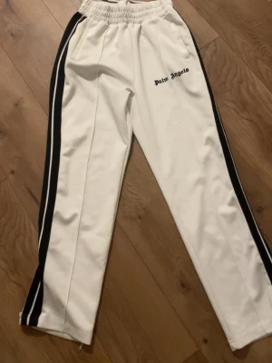 Vita mjukisbyxor Palm Angels - Säljer ett par vita mjukisbyxor från Palm Angels med svarta sidoränder och elastisk midja. Byxorna har en avslappnad passform och loggan broderad på vänster sida. Perfekta för en chill och trendig look.