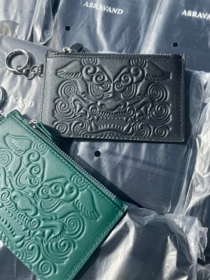 Svart och grön plånbok från asravand (bundle) - Säljer dessa supersnygga korthållare/plånböcker med unikt präglat motiv från märket Asravand.                                                                   De är helt nya och tillverkade i högkvalitativt, äkta läder.Varje korthållare har en smidig dragkedja samt en medföljande rem/band så att du enkelt kan bära den runt halsen.Färger: Svart och grön.Material: 100% äkta läder. Tillbehör: Avtagbart band för halsen ingår.Skick: Helt nya / oanvända.