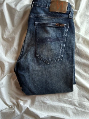 Mörkblå jeans från Nudie Jeans - Säljer ett par mörkblå jeans från Nudie Jeans med klassisk femficksmodell och snygga slitningar framtill. Jeansen har en skön passform och är tillverkade i denim med tydliga kontrastsömmar och läderpatch bak i midjan. Model skinny lin.