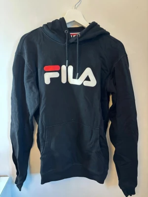 Svart hoodie från FILA  - Säljer en svart hoodie från FILA med stor vit och röd logga på bröstet. Hoodien har huva och en stor magficka framtill. Tillverkad i mjukt material som passar perfekt för en avslappnad stil.