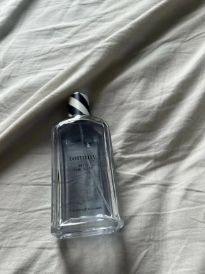 Tommy Hilfiger Into The Surf parfym - Fräsch parfym från Tommy Hilfiger, 'Into The Surf', i en genomskinlig glasflaska med blått och vitt randigt lock. Flaskan har en stilren, rektangulär form och en modern design som passar perfekt för dig som gillar sportiga och fräscha dofter.