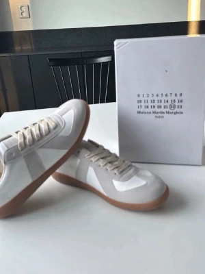 Maison Margiela vita sneakers - Säljer ett par stilrena vita sneakers från Maison Margiela med beige mockadetaljer och gummisula. Skorna har klassisk låg profil, vita skosnören och en snygg retrovibe. Perfekta för dig som gillar minimalistisk och trendig design.